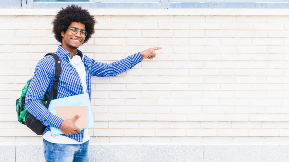 portrait-african-smiling-male-student-holding-books-hand-pointing-finger-white-painted-wall